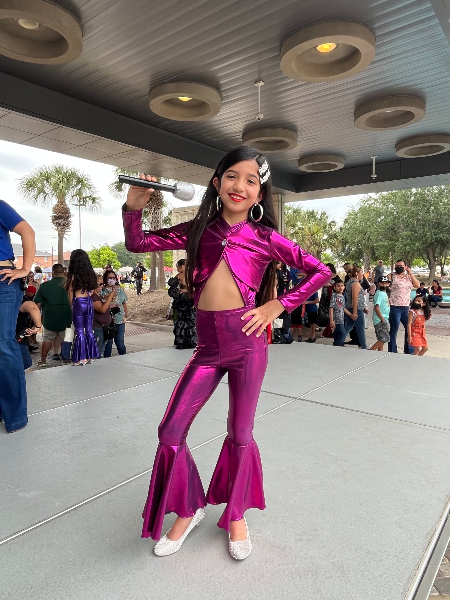Child dressed as Selena poses on stage with a microphone during a costume contest at Main Library.